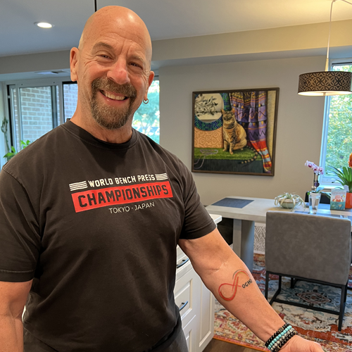  Howard Aaron, wearing a World Bench Press Championships t-shirt, shows a tattoo of a red infinity sign with the word Ocho next to it on his arm.